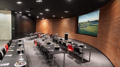 Große Leinwand, Rednerpult, Konferenz-Bestuhlung mit Tisch in Reihen. Raum mit Holz modern vertäfelt.