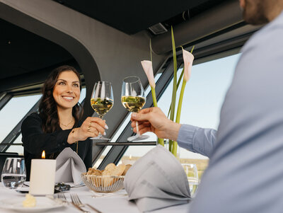 Eine Frau sitzt einem Mann gegenüber. Sie prosten sich mit einem Glas Weißwein zu.