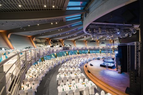 Porsche Auditorium - Mischbestuhlung (inkl. Galerie): bis zu 800 Personen Abgerundeter Raum. Historische Fahrzeuggalerie offen über dem Auditorium. Galabestuhlung, Bankett, auch historische Fahrzeugausstellung bestuhlt.. Bühne mit großer Leinwand und Rednerpult. Austellungsfahrzeug auf der Bühne. Im Hintergrund Panoramafenster mit Blick auf die Rundstrecke.