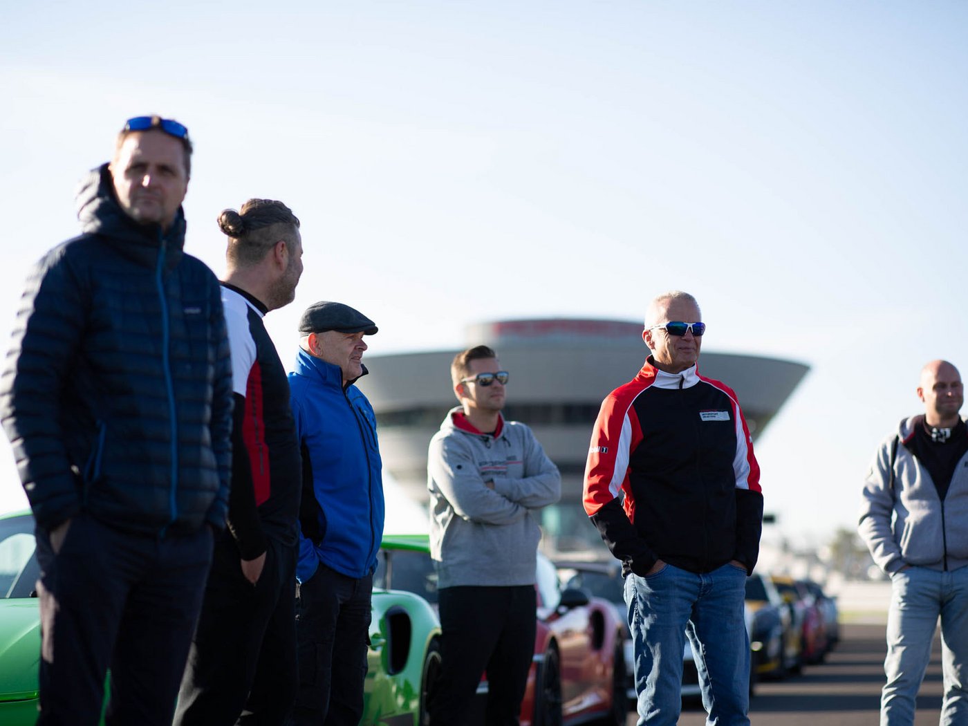 Welche Personen stehen neben ihren Fahrzeugen auf der Rennstrecke des Porsche experience Center in Leipzig und werden von einem Instrukteur eingewiesen