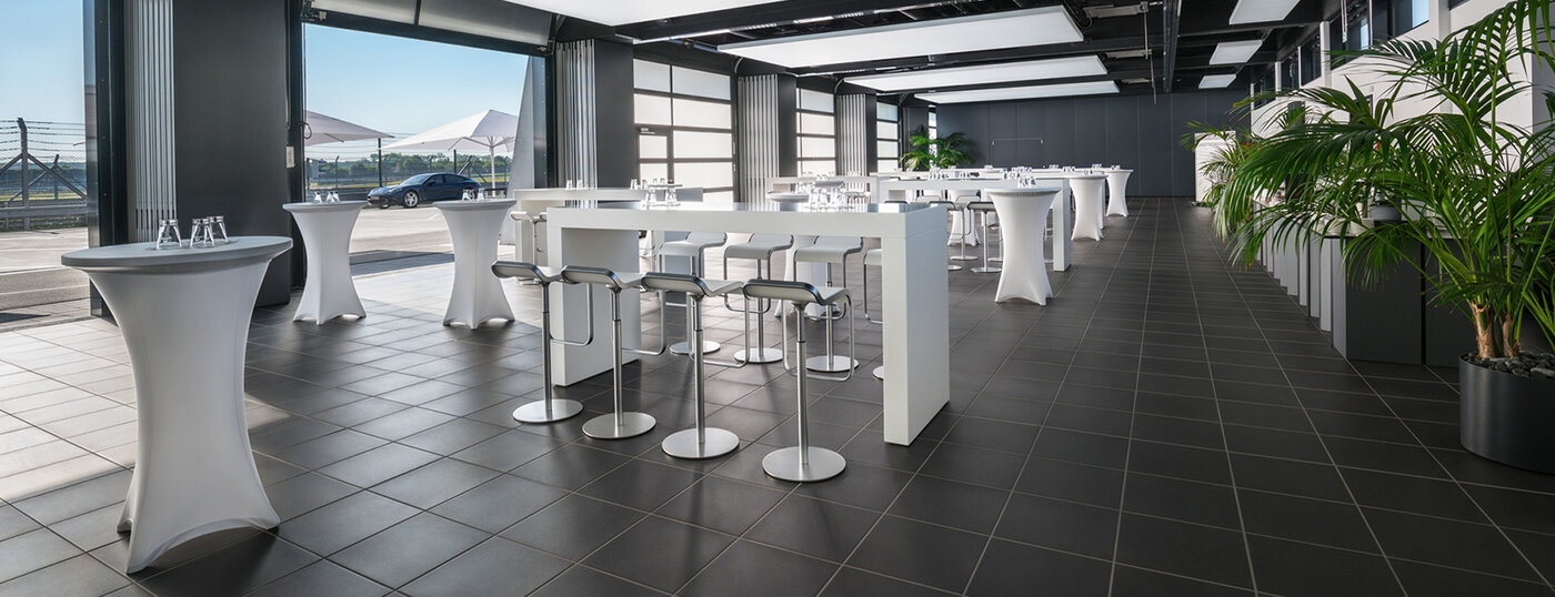 Large hall, bar tables and bar stools. Bar. Roller doors open. Terrace built into the pit lane.