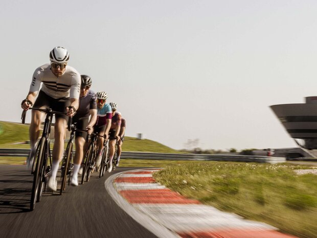 5 Rennradfahrer fahren in einer Reihe über die Rundstrecke. Sie tragen Fahrradtrikots, Sonnenbrille und Helm.