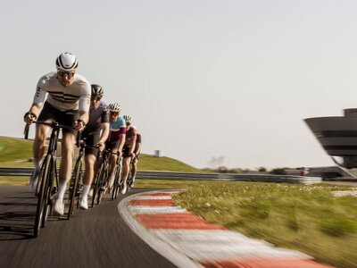 5 Rennradfahrer fahren in einer Reihe über die Rundstrecke. Sie tragen Fahrradtrikots, Sonnenbrille und Helm.
