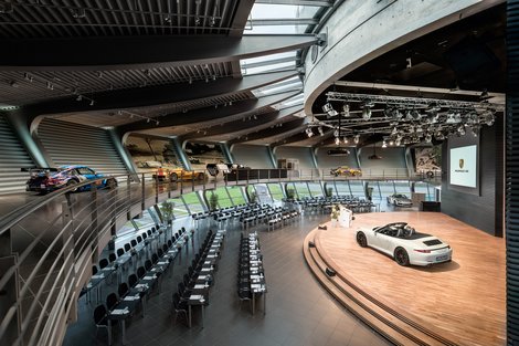 Porsche Auditorium - Konferenzbestuhlungen Parlamentarisch: bis zu 200 Personen Abgerundeter Raum. Historische Fahrzeuggalerie offen über dem Auditorium. Konferenzbestuhlung mit Tisch in Reihen. Bühne mit großer Leinwand und Rednerpult. Austellungsfahrzeug auf der Bühne. Im Hintergrund Panoramafenster mit Blick auf die Rundstrecke.