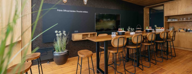 Raum mit Holzvertäfelung, Outdoor Look, kleine Teeküche, großer Tich in der Mitte mit 12 Hochstühlen.