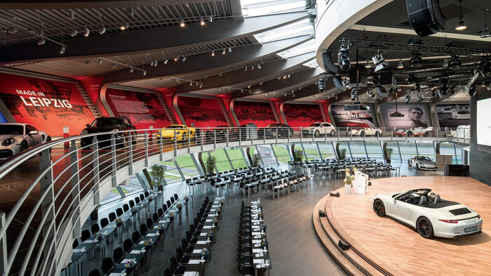 A hall can be seen. It is round and has large windows. A Porsche 911 stands on a wooden platform. Conference chairs are set up in several rows in front of the platform. Above the hall, a gallery can be seen displaying a historical Porsche vehicle exhibition.