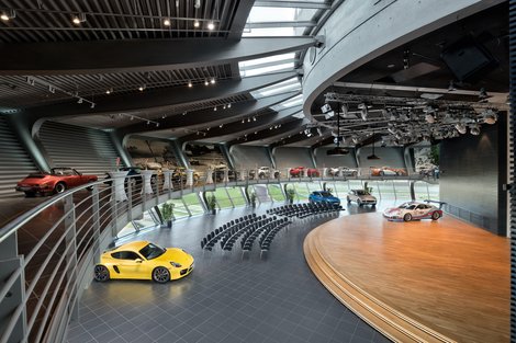 Porsche Auditorium - Konferenzbestuhlungen Reihe: bis zu 500 Personen Abgerundeter Raum. Historische Fahrzeuggalerie offen über dem Auditorium. Konferenzbestuhlung. Bühne mit großer Leinwand und Rednerpult. Austellungsfahrzeug auf der Bühne. Im Hintergrund Panoramafenster mit Blick auf die Rundstrecke.