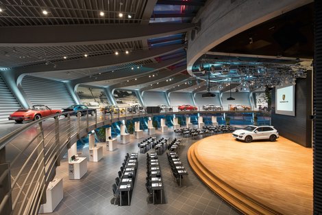 Porsche Auditorium - Konferenzbestuhlungen Parlamentarisch: bis zu 200 Personen Abgerundeter Raum. Historische Fahrzeuggalerie offen über dem Auditorium. Konferenzbestuhlung mit Tisch in Reihen. Aufgebaute Stände im Hintergrund. Bühne mit großer Leinwand und Rednerpult. Austellungsfahrzeug auf der Bühne. Im Hintergrund Panoramafenster mit Blick auf die Rundstrecke.
