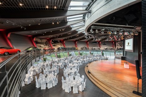 Porsche Auditorium - Galabestuhlungen Bankett: bis zu 330 Personen Abgerundeter Raum. Historische Fahrzeuggalerie offen über dem Auditorium. Galabestuhlung, runde Tische. Bühne mit großer Leinwand und Rednerpult. Austellungsfahrzeug auf der Bühne. Im Hintergrund Panoramafenster mit Blick auf die Rundstrecke.