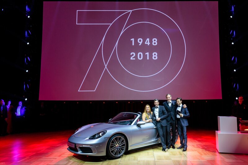 Die Spendentombolagewinner stehen vor dem Haupgewinn, ein Porsche 718 Boxster. Dahinter eine Leinwand mit 70 Jahre Porsche Sportwagen.