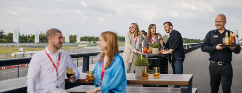 mehrer junge Personen genießen auf der Terasse des Carrera Cafes Getränke. Es sind Stehtische und Tische mit Sitzmöglichekiten zu sehen. Eine Bedienung bringt Getränke raus.