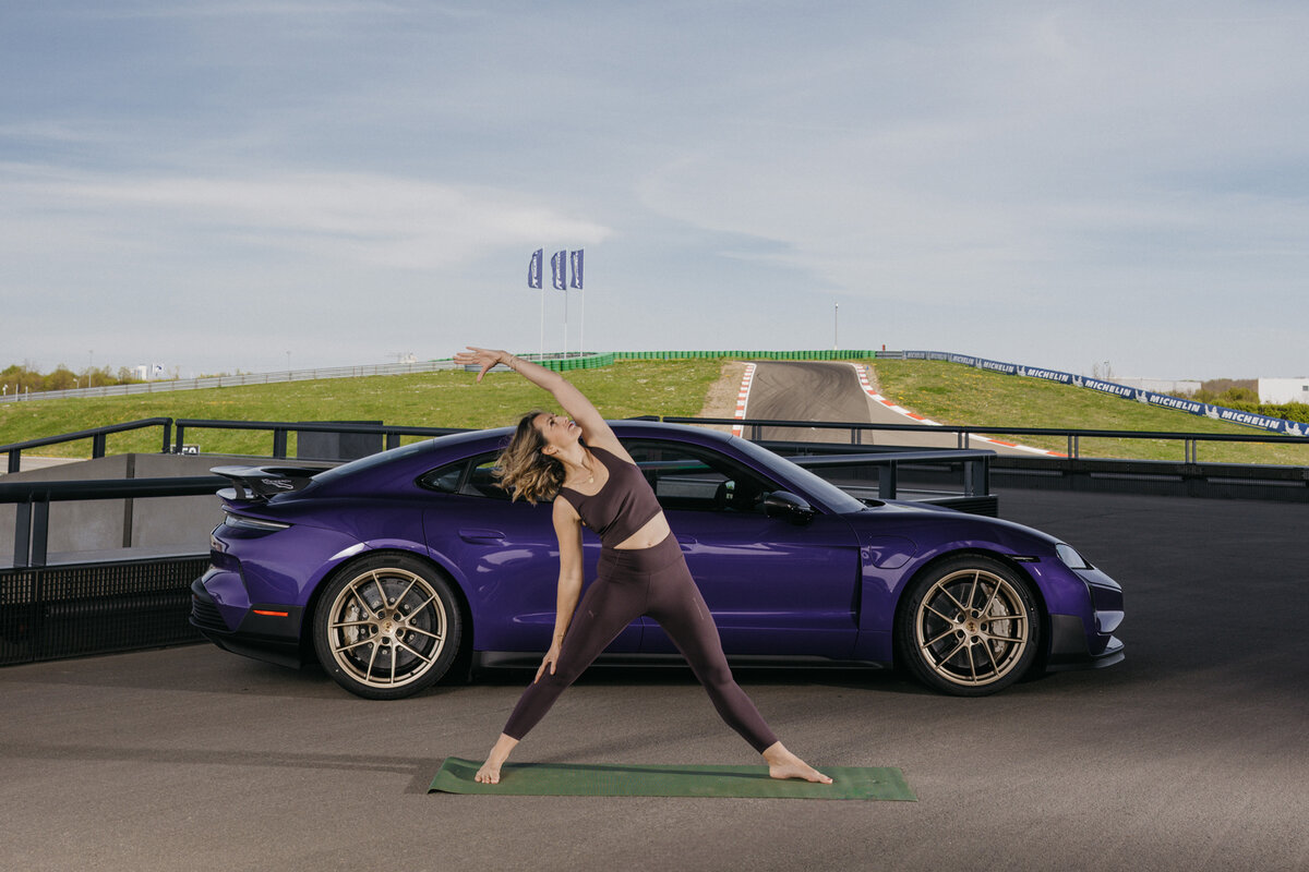 Eine junge Frau in Sportklamotten macht eine Yogaübung auf der terasse vor dem Carrera Cafe. Hinter ihr steht ien Porsche Taycan. Im Hintergrund ist die Rundstrecke zu sehen. 