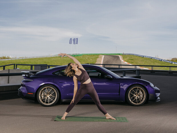 Eine junge Frau in Sportklamotten macht eine Yogaübung auf einer Matte. Hinter ihr steht ein Porsche Taycan. Im Hintergrund ist die Runstrecke zu sehen.