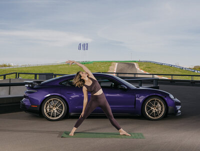 Eine junge Frau in Sportklamotten macht eine Yogaübung auf einer Matte. Hinter ihr steht ein Porsche Taycan. Im Hintergrund ist die Runstrecke zu sehen. 
