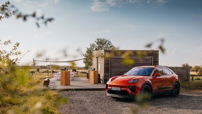 Ein Cayenne steht vor dem Off-Road Gelände bei Sonnenuntergang. Es ist viel Natur zu sehen und eine moderne Holzhütte, die Treffpunkt für das Off-Road Gelände ist.