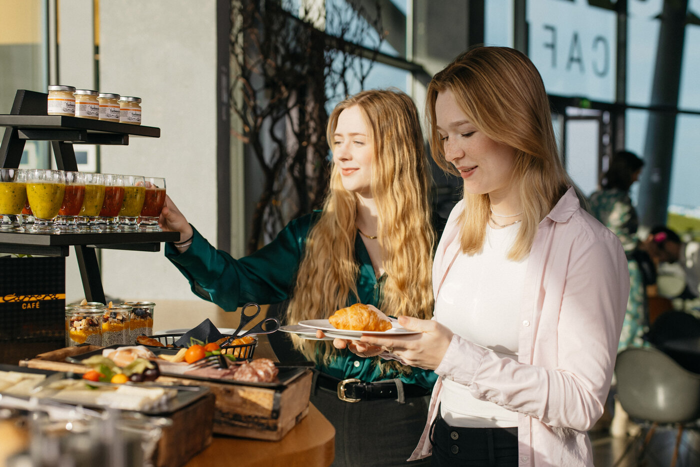 zwei junge Frauen bedienen sich am Früstücksbuffet. Es gibt diverse Smoothise und Säfte. Eine Käseplatte und diverse Schinken und Salami. Zusätzlich Obst und Chia-Pudding. 