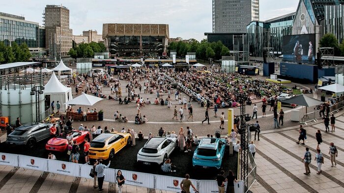 Es ist der Augustusplatz zu sehen. Rechts das Augusteum der Universität Leipzig. Geradeaus das Gewandhaus Leipzigs. Vorne ist ein Podest zu sehen auf dem verschiedene Porsche Modelle zu sehen sind. Vor dem Augusteum ist eine Leinwand aufgebaut, die ein Stück aus der Oper zeigt. davor sind Sitzmöglichekiten. Es sind ganz viele Menschen auf dem Platz und bei der Autoausstellung.
