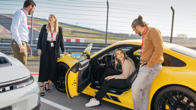 Three people are standing around a 911, one person is sitting inside. The driver's door is open. The car is in the pit lane and ready for the driving program.
