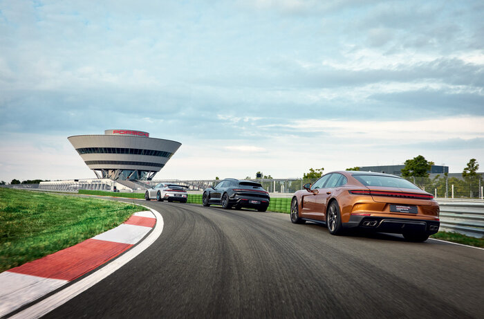 Bild Fahrerlebnis. Zwei Porsche Panamera und ein Taycan fahren hintereinander auf der Rundstrecke. Im Hintergrund ist das Experience Center zu sehen.