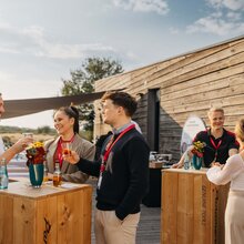 Several people are standing around high wooden tables enjoying drinks.
