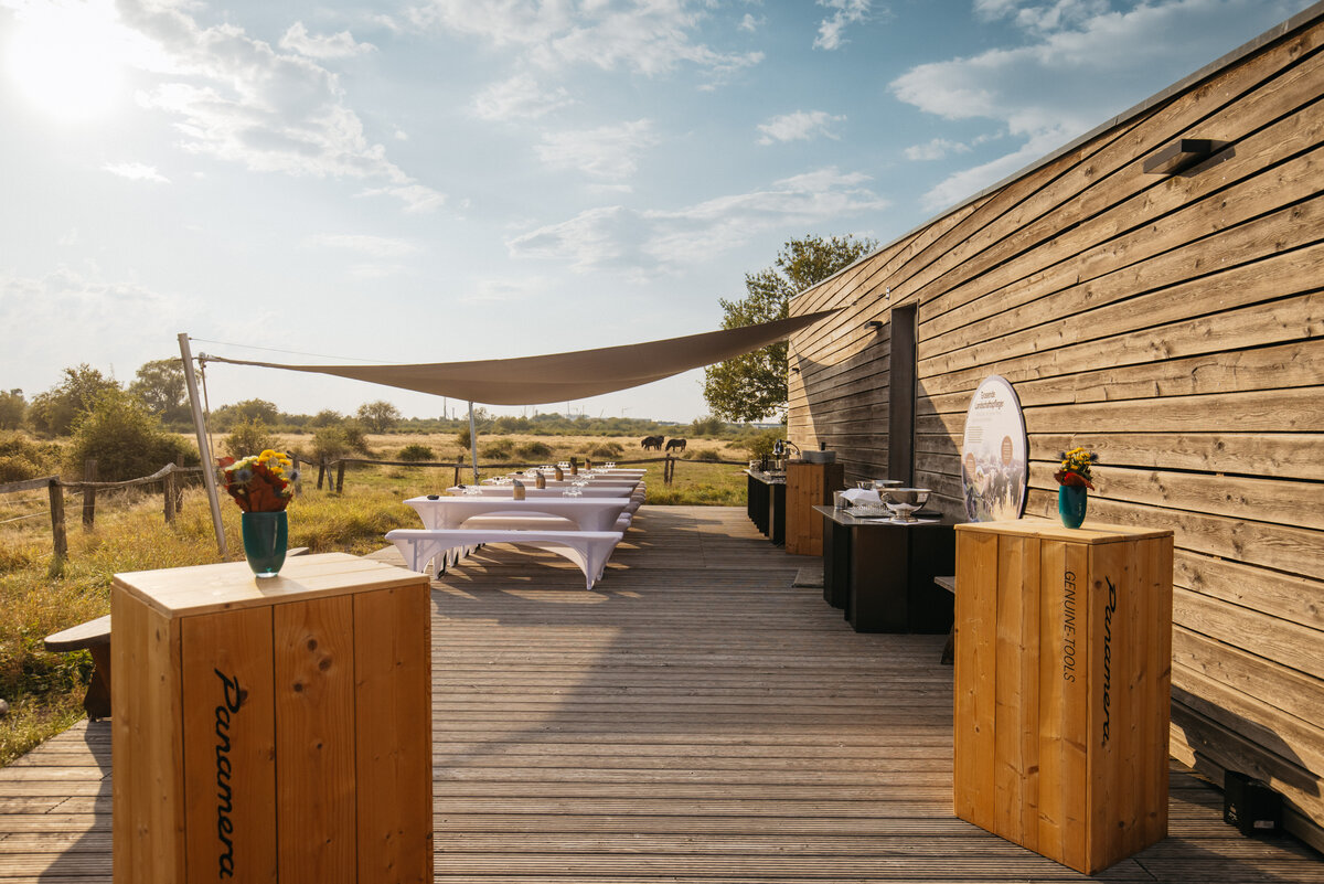 Wooden terrace with square wooden bar tables. Behind them are several rows of beer benches with elegant covers. To the right is the bar and catering area. Sun sails provide protection from the sun.