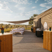 Wooden terrace with square wooden bar tables. Behind them are several rows of beer benches with elegant covers. To the right is the bar and catering area. Sun sails provide protection from the sun.