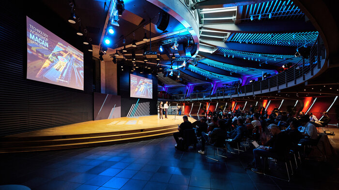 A darkened auditorium can be seen. Two people are standing on the illuminated stage giving a speech. Many people are sitting in front of the stage in several rows, watching.