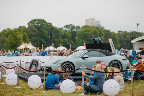 Auf einem Podest ist ein Porsche 911 cabrio. Darin sitzen zwei Personen, die dem Konzert zuhören. Um sie herum sitzen viele Besucher auf Picknickdecken und Campingstühlen und hören auch dem Konzert zu. 