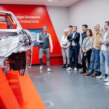 Eine Besuchergruppe steht in einem Ausstellungsraum. An den Wänden sind Bildschirme, die dei unterschiedlichen Schritte des karosseriebaus erklären. In der Mitte steht eine karosserie als Beispiel. Der Werksführer erklärt was man sieht.