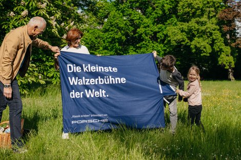 Großeltern und ihre beiden Enkel breiten eine Picknickdecke auf einer Wiese aus. 