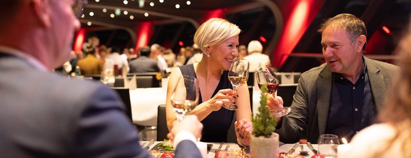 Ein schick gekleidetes älteres Paar stoßt mit Wein an. Sie sitzen im Restaurant an einem festlich gedeckten Tisch mit weiteren Kollegen.