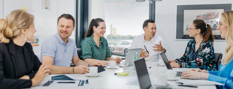 Eine Gruppe Büroangestellte sitzt an einem Tisch und hält ein Meeting ab. Der Raum ist modern und hat ein großes Fenster. 